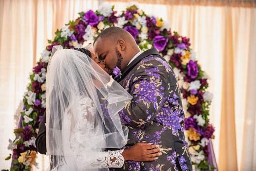 Moses and Crystal kissing at their wedding under an arch of purple and white flowers